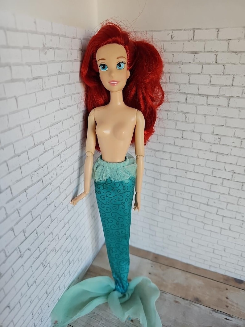 May include: A doll of a mermaid with red hair and blue eyes. The doll has a teal tail with a swirling pattern and a matching teal top. The doll is standing in front of a white brick wall.