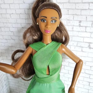 May include: A fashion doll with brown hair in a high ponytail and flowing waves. The doll has brown skin, blue eyes, and wears a green dress with a crisscross bodice. The doll is accessorized with small gold earrings.