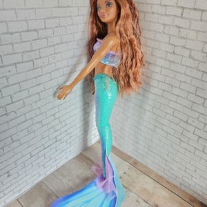 May include: A mermaid doll with long, auburn hair and a smiling expression. The doll wears a sparkly, two-tone top and a turquoise mermaid tail. The tail has a large, blue fin with purple accents. The doll is standing on a light blue base.
