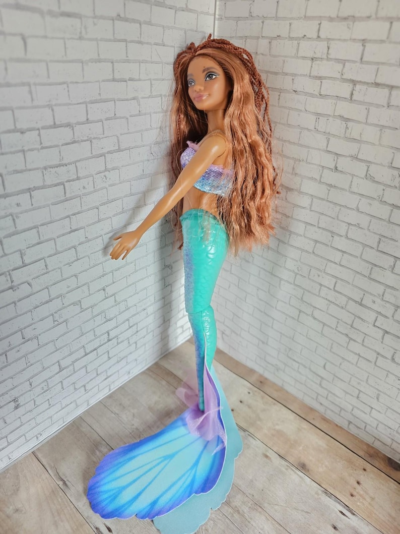 May include: A mermaid doll with long, auburn hair and a smiling expression. The doll wears a sparkly, two-tone top and a turquoise mermaid tail. The tail has a large, blue fin with purple accents. The doll is standing on a light blue base.