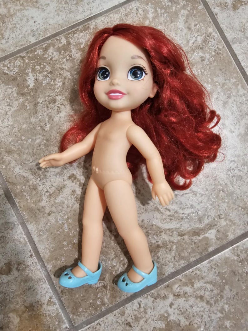 May include: A doll with red hair, blue eyes, and light blue shoes. The doll has a nude-colored body and is posed with arms outstretched. The doll's hair is styled in loose waves. The shoes have a strap and a heart-shaped cutout.