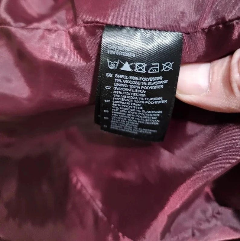 Puede incluir: Primer plano de una etiqueta de ropa negra con texto blanco. La etiqueta enumera la composici&oacute;n de la prenda, incluido el forro y otros materiales. La etiqueta se sostiene contra una tela burdeos.