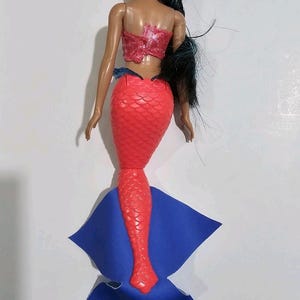 Puede incluir: Una mu&ntilde;eca de sirena con cabello negro y un top rojo con lentejuelas. La mu&ntilde;eca tiene una cola roja escamada y una aleta azul. La mu&ntilde;eca es un juguete y probablemente est&aacute; hecha de pl&aacute;stico.