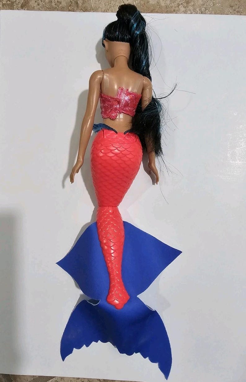 Puede incluir: Una mu&ntilde;eca de sirena con cabello negro y un top rojo con lentejuelas. La mu&ntilde;eca tiene una cola roja escamada y una aleta azul. La mu&ntilde;eca es un juguete y probablemente est&aacute; hecha de pl&aacute;stico.