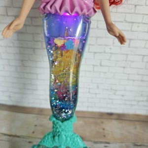 Peut inclure: Poup&eacute;e Ariel avec des cheveux roux et un haut rose. La queue de la poup&eacute;e est un r&eacute;cipient transparent rempli de paillettes avec une sc&egrave;ne de ch&acirc;teau et une base turquoise. La queue s'illumine d'une lueur violette.
