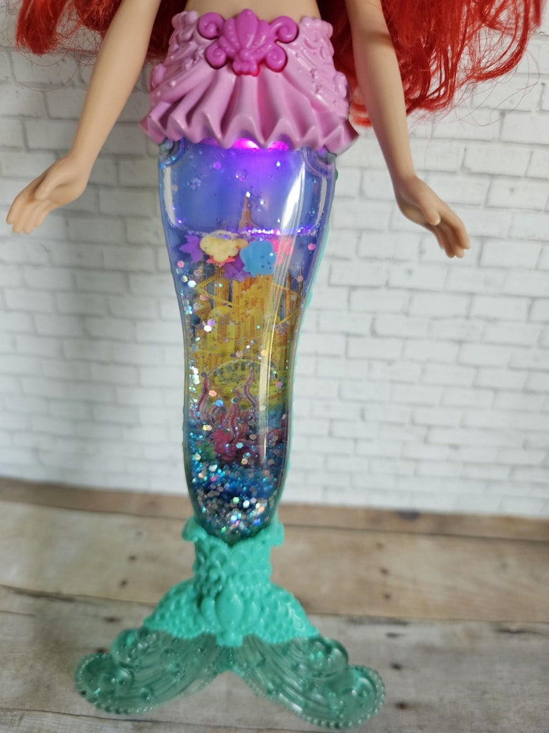 Peut inclure: Poup&eacute;e Ariel avec des cheveux roux et un haut rose. La queue de la poup&eacute;e est un r&eacute;cipient transparent rempli de paillettes avec une sc&egrave;ne de ch&acirc;teau et une base turquoise. La queue s'illumine d'une lueur violette.