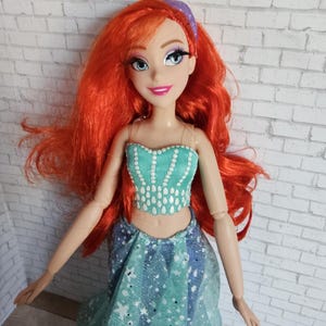 May include: A doll with vibrant red hair and a teal top and skirt. The doll has a purple headband and is smiling. The top is decorated with white teardrop shapes. The skirt is blue with white star shapes.