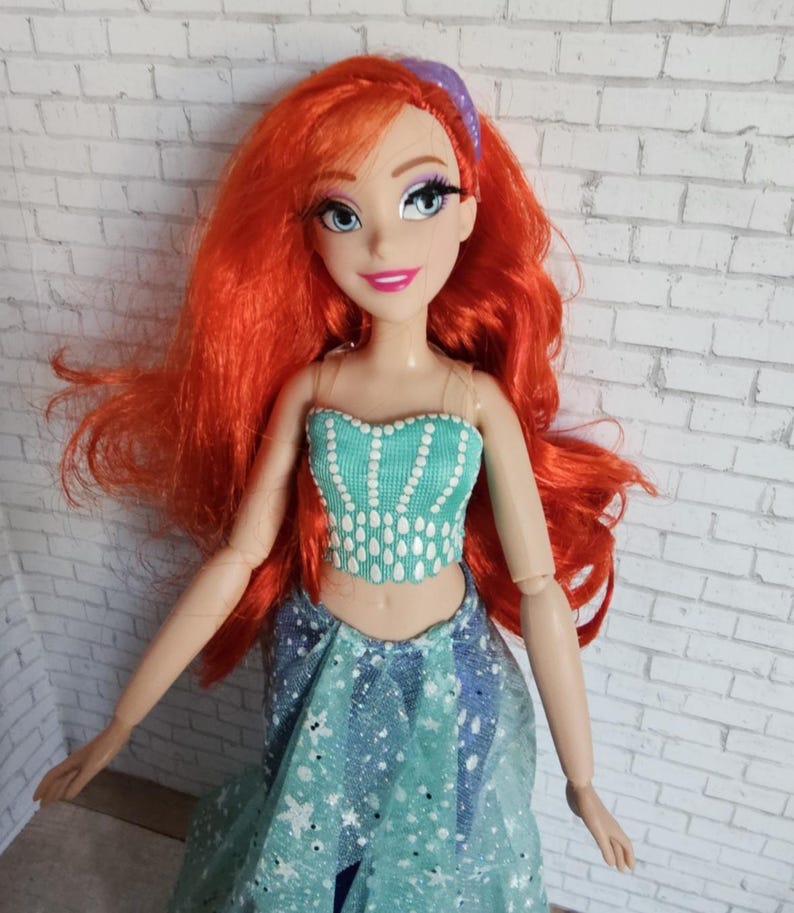 May include: A doll with vibrant red hair and a teal top and skirt. The doll has a purple headband and is smiling. The top is decorated with white teardrop shapes. The skirt is blue with white star shapes.