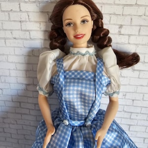 Puede incluir: Muñeca con cabello castaño trenzado, vestida con un vestido a cuadros azul y blanco con mangas blancas. La muñeca tiene una blusa blanca con ribete azul y lápiz labial rojo. Objeto de colección.