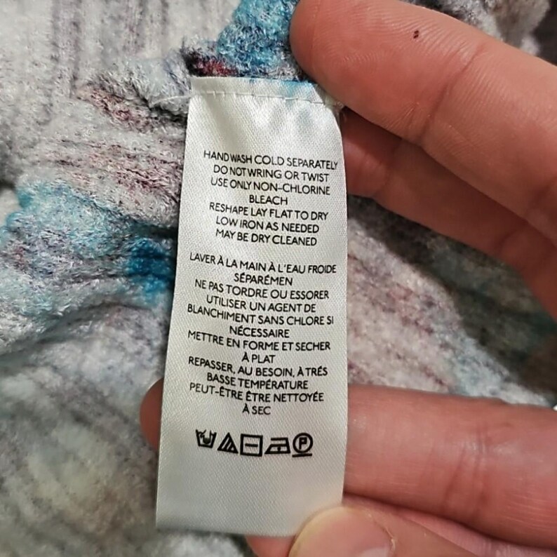 Puede incluir: Una etiqueta de ropa blanca con texto negro que detalla las instrucciones de cuidado. La etiqueta incluye pautas de lavado, secado y planchado. La tela de fondo es una mezcla de azul, gris y granate.