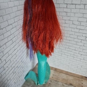May include: A doll with long red hair, a purple streak, and a turquoise mermaid tail. The doll is positioned against a white brick wall and a wooden surface.