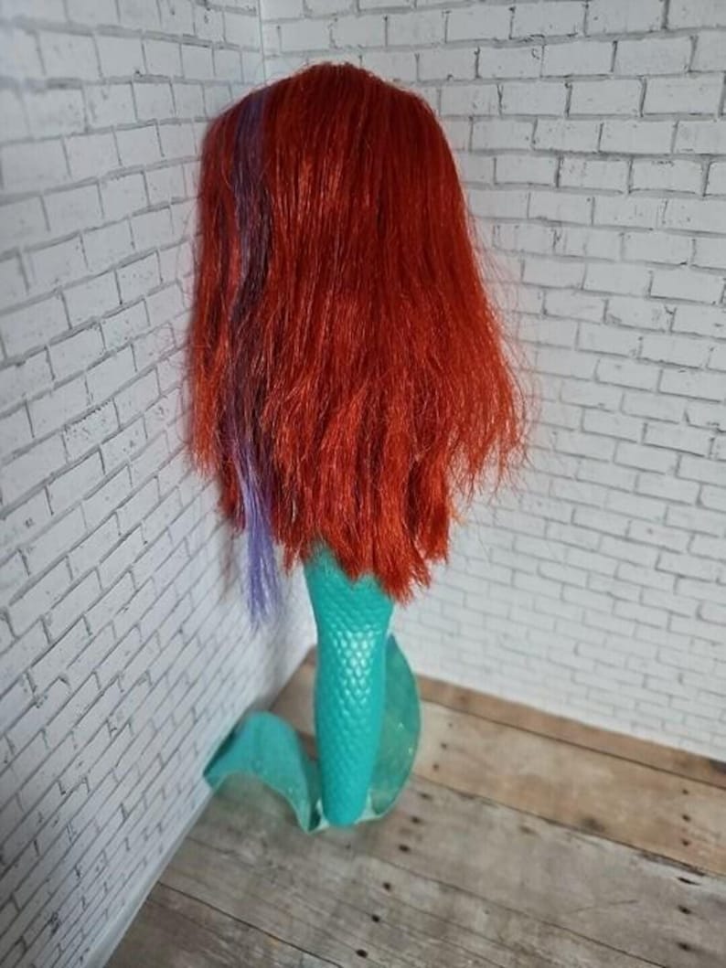 May include: A doll with long red hair, a purple streak, and a turquoise mermaid tail. The doll is positioned against a white brick wall and a wooden surface.