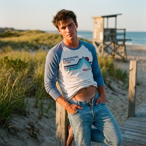May include: A light grey and blue raglan-sleeve shirt with the text "Abercrombie & Fitch Co. Est. 1892 New York" and a mountain graphic. The shirt is paired with light blue jeans. The background includes a beach, dunes, and a lifeguard stand.