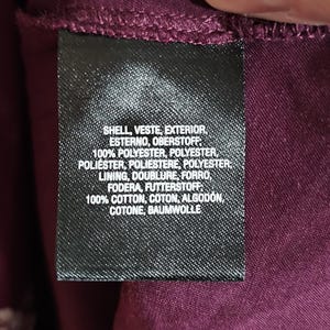 Pu&ograve; includere: Primo piano di un'etichetta di abbigliamento nera con testo bianco. L'etichetta elenca i materiali e le loro percentuali, tra cui poliestere e cotone. Lo sfondo &egrave; un tessuto bordeaux scuro.