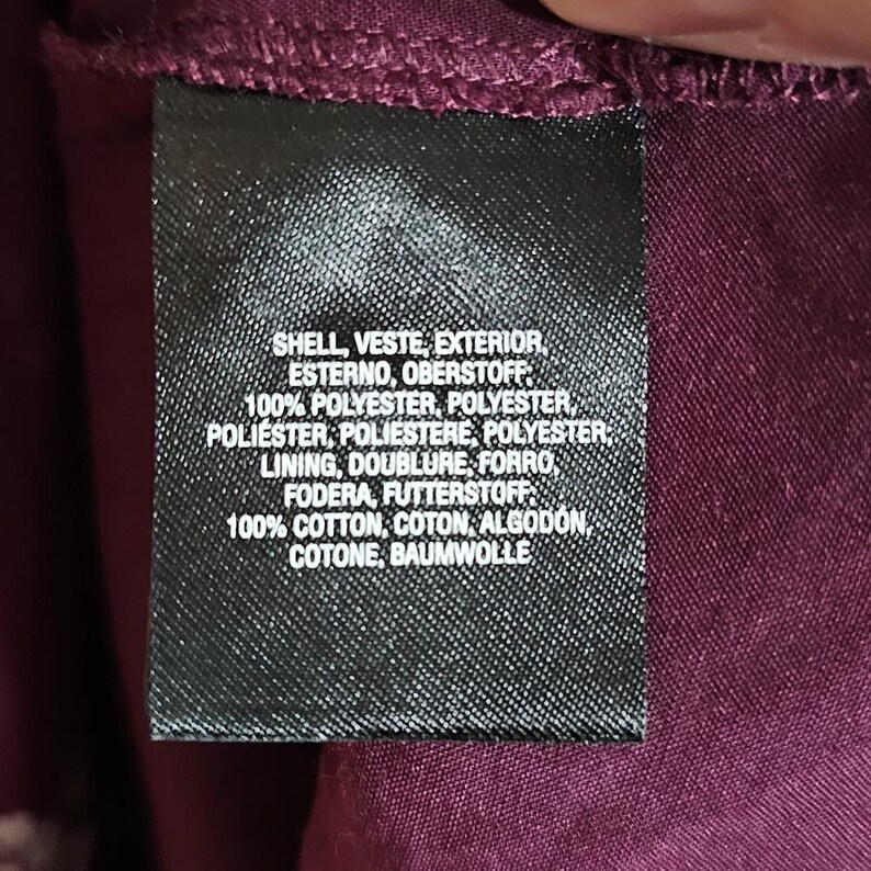 Pu&ograve; includere: Primo piano di un'etichetta di abbigliamento nera con testo bianco. L'etichetta elenca i materiali e le loro percentuali, tra cui poliestere e cotone. Lo sfondo &egrave; un tessuto bordeaux scuro.