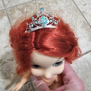 May include: A doll with red hair and a silver tiara with blue accents. The tiara is shaped like a seashell and has small blue gems. The doll's face is pale with blue eyes and a slight smile.