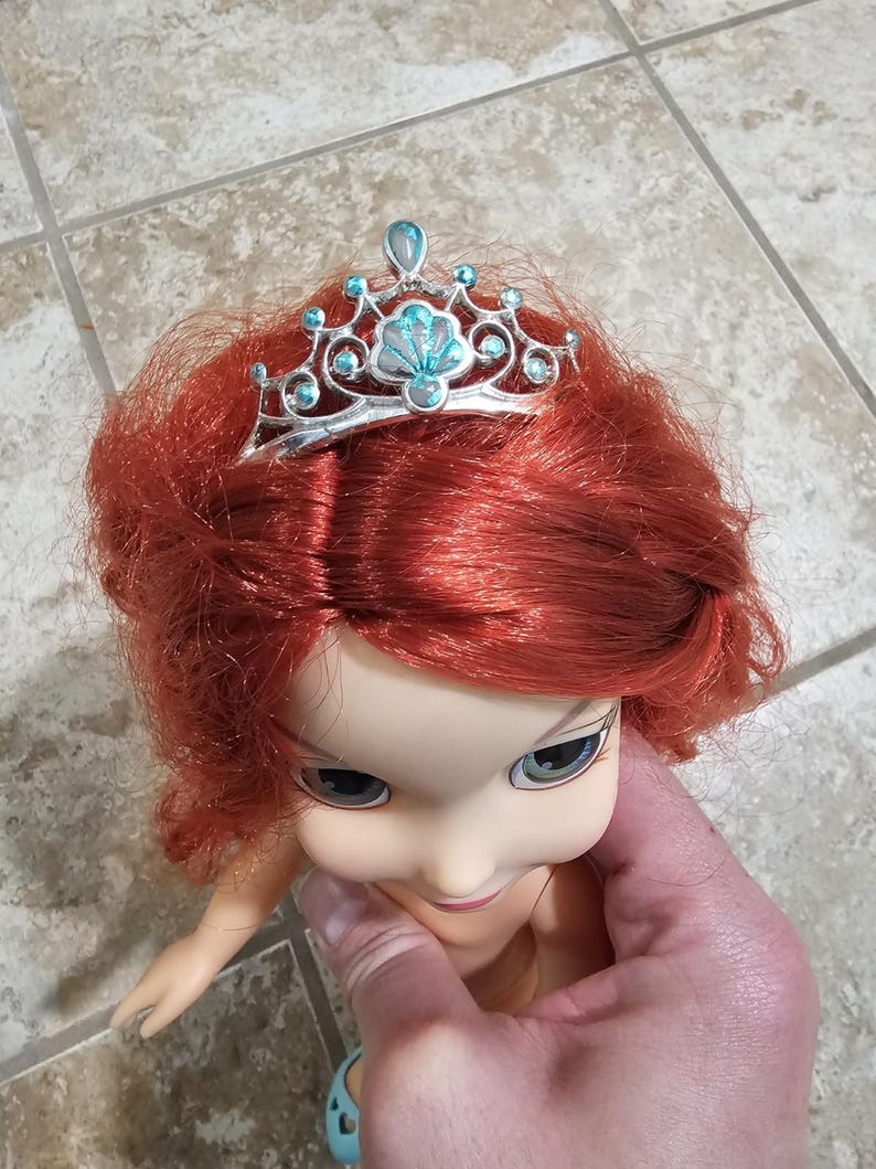 May include: A doll with red hair and a silver tiara with blue accents. The tiara is shaped like a seashell and has small blue gems. The doll's face is pale with blue eyes and a slight smile.