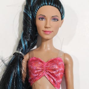 Puede incluir: Mu&ntilde;eca con pelo largo negro con mechones azules, peinado en una cola de caballo alta. La mu&ntilde;eca tiene tez bronceada, l&aacute;piz labial rosa y un top de lentejuelas rosa y naranja. Los ojos de la mu&ntilde;eca son de color marr&oacute;n claro.