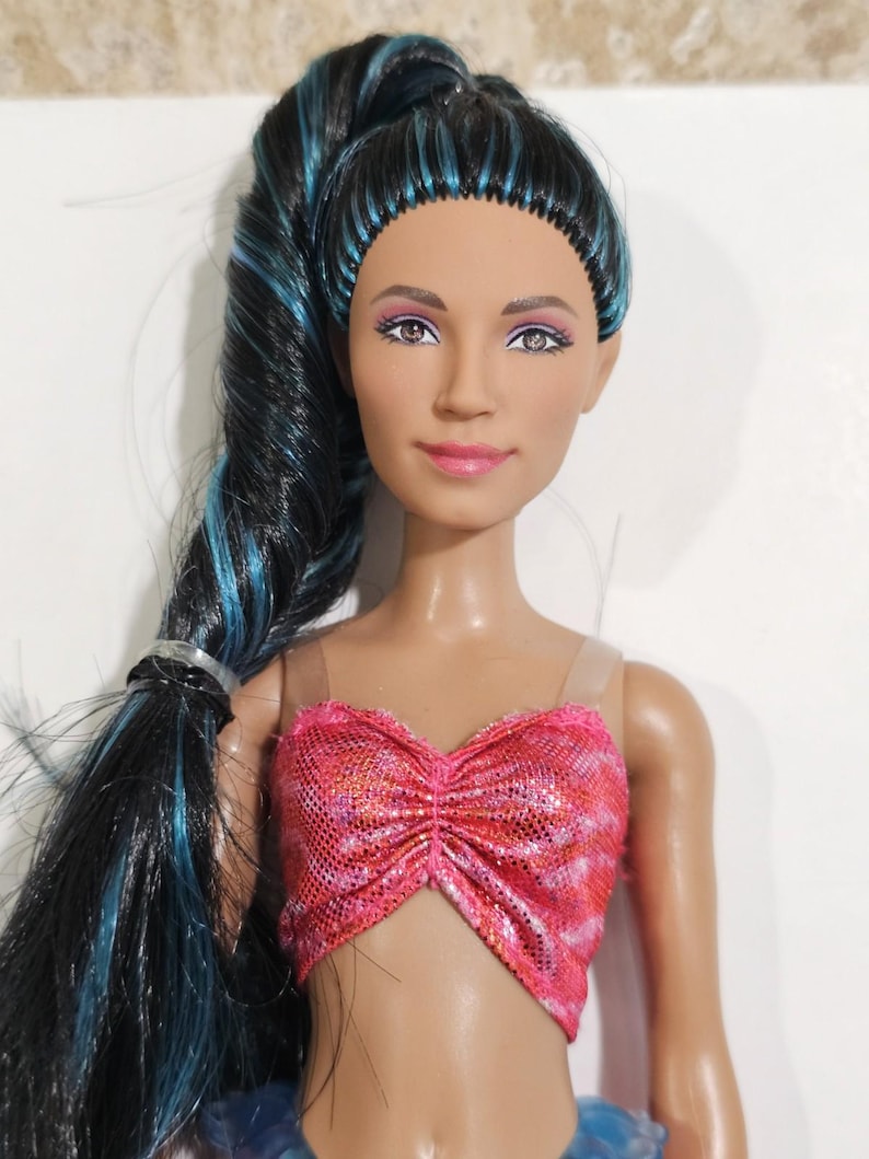 Puede incluir: Mu&ntilde;eca con pelo largo negro con mechones azules, peinado en una cola de caballo alta. La mu&ntilde;eca tiene tez bronceada, l&aacute;piz labial rosa y un top de lentejuelas rosa y naranja. Los ojos de la mu&ntilde;eca son de color marr&oacute;n claro.