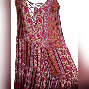 May include: A sleeveless, tiered dress with a floral pattern in shades of pink, beige, and brown. The dress features a lace-up neckline and a flowing, bohemian style. The fabric has a soft, textured appearance.