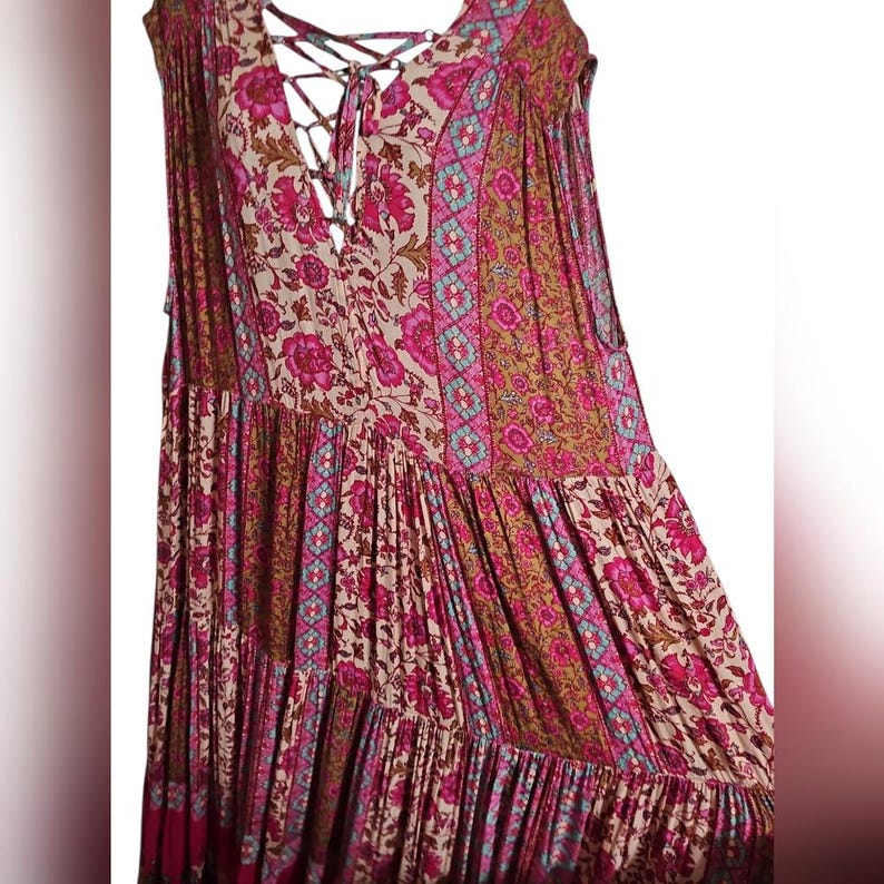 May include: A sleeveless, tiered dress with a floral pattern in shades of pink, beige, and brown. The dress features a lace-up neckline and a flowing, bohemian style. The fabric has a soft, textured appearance.