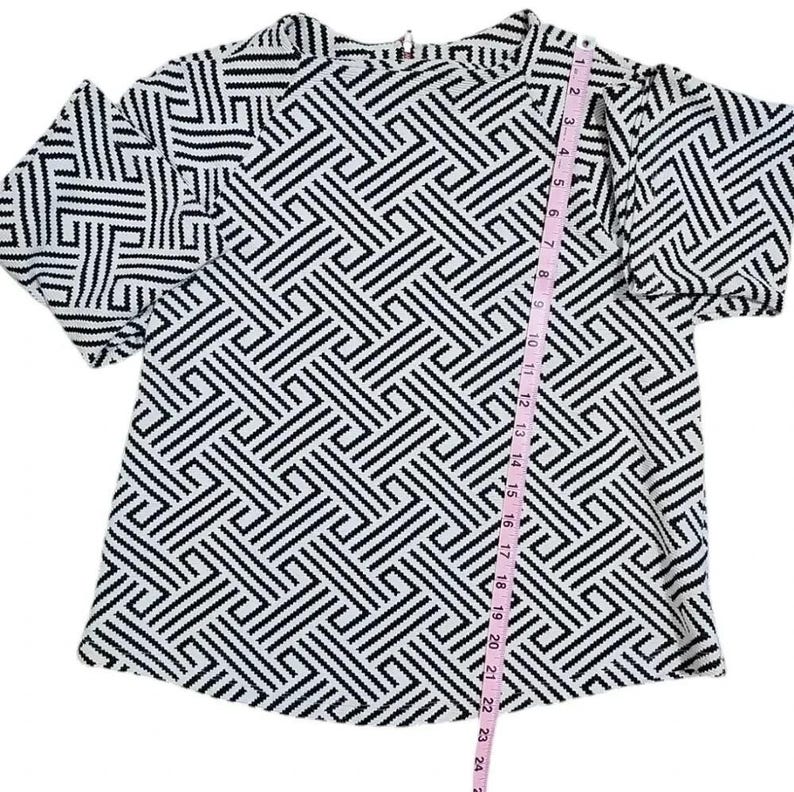 May include: A black and white geometric patterned top with short sleeves. The top has a crew neck and a zipper closure at the back. A pink measuring tape is placed vertically along the front of the top.