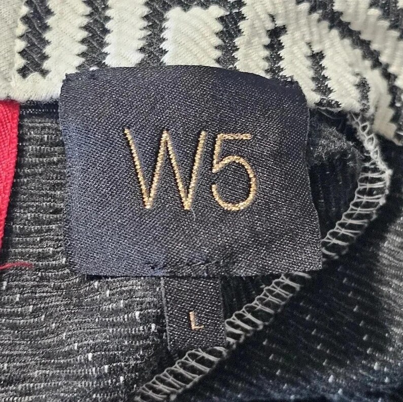 May include: Close-up of a black fabric tag with the gold embroidered text "W5" and the size "L". The tag is sewn onto a textured black and white fabric. A red fabric is visible on the left side.