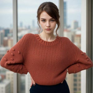 Puede incluir: Un suéter de punto color óxido con cuello redondo y detalles de bolitas texturizadas en las mangas. El suéter lo lleva una persona con las manos en las caderas. Se ve un paisaje urbano a través de una ventana.