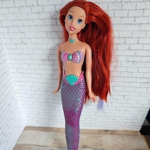May include: A doll of a mermaid with long red hair, blue eyes, and a pink and teal seashell top and tail. The doll has a teal necklace and a pink seashell belt. The mermaid doll is standing in front of a white brick wall.