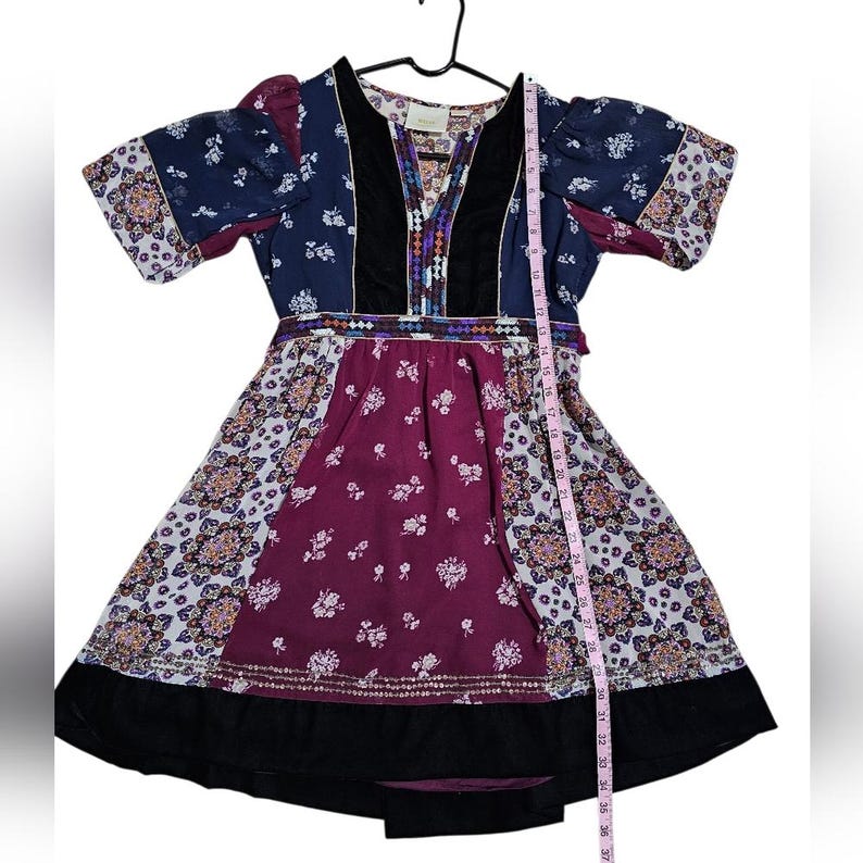 Pu&ograve; includere: Un abito colorato con scollo a V e maniche corte. L'abito presenta un mix di fantasie, tra cui disegni floreali e geometrici, nelle tonalit&agrave; del blu, viola e bianco. L'abito ha un orlo nero ed &egrave; lungo circa 94 cm.