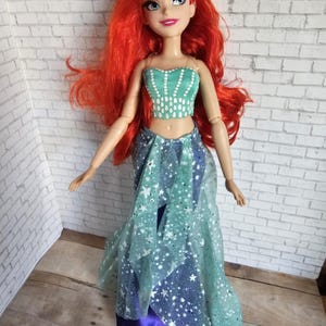 May include: A doll with flowing red hair, dressed in a teal crop top and a layered skirt with a blue and white star pattern. The doll has a purple headband, set against a white brick wall.
