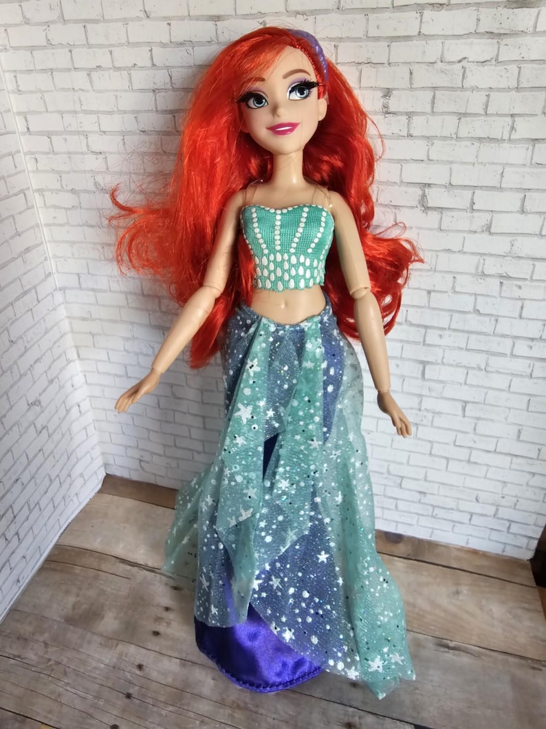 May include: A doll with flowing red hair, dressed in a teal crop top and a layered skirt with a blue and white star pattern. The doll has a purple headband, set against a white brick wall.