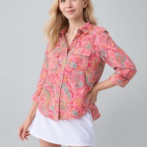 May include: A pink button-down shirt with a paisley pattern in shades of blue, yellow, and green. The shirt has two chest pockets and three-quarter sleeves. The person is wearing a white skirt.