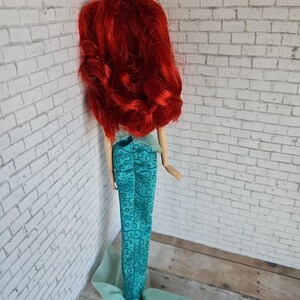 May include: A doll with long, vibrant red hair and a teal mermaid tail. The tail features a swirling pattern and a flowing, sheer fabric at the bottom. The doll is positioned against a white brick wall.