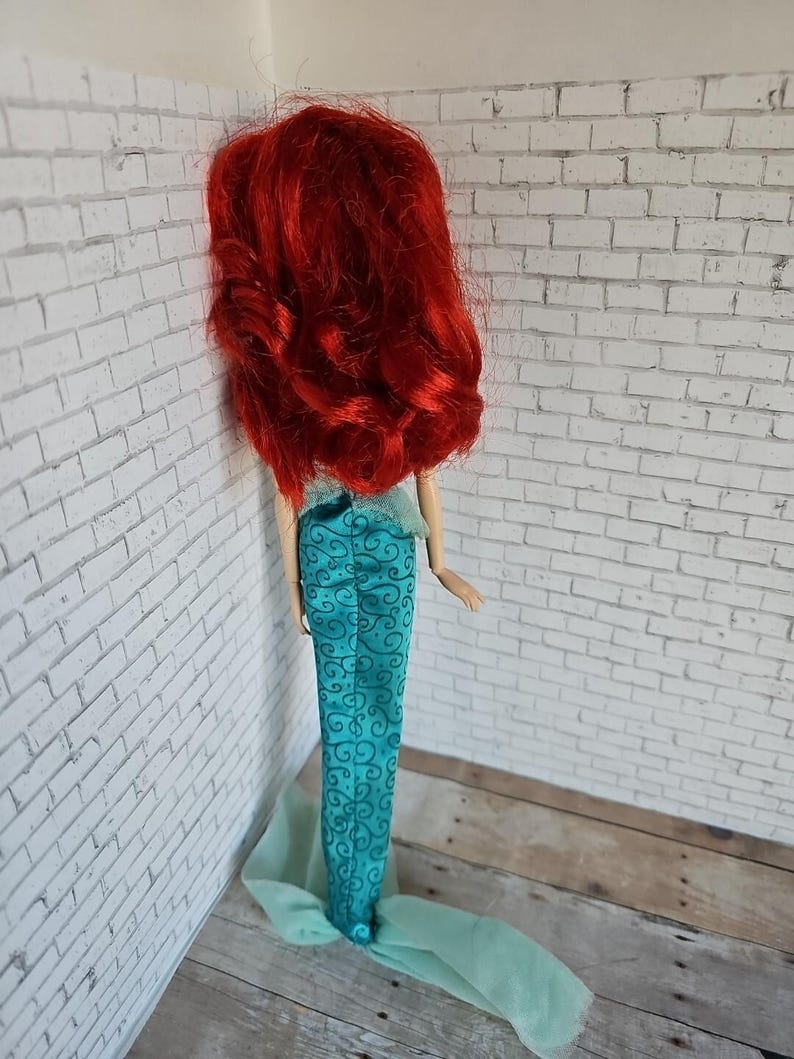 May include: A doll with long, vibrant red hair and a teal mermaid tail. The tail features a swirling pattern and a flowing, sheer fabric at the bottom. The doll is positioned against a white brick wall.