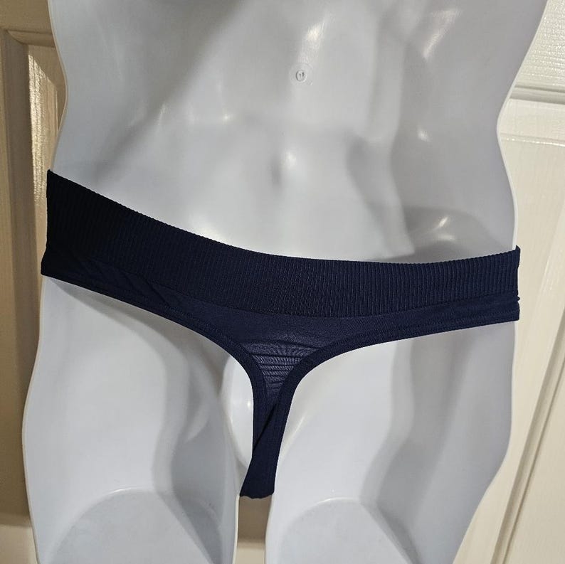 K&ouml;nnte beinhalten: Marineblauer String-Tanga mit geripptem Bund. Der String hat vorne einen durchsichtigen, netzartigen Stoff und einen d&uuml;nnen Riemen. Die Unterw&auml;sche wird auf einer wei&szlig;en Schaufensterpuppe pr&auml;sentiert.