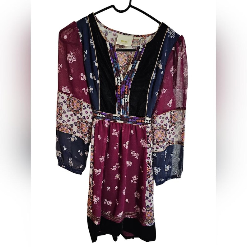 Pu&ograve; includere: Abito a maniche lunghe con scollo a V e un mix di fantasie e colori. L'abito presenta un pannello in velluto nero sul davanti, con motivi floreali e geometrici nei toni del bordeaux, blu navy e crema. Appeso a una gruccia nera.