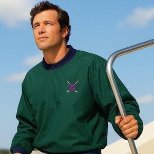 May include: A dark green golf pullover with a navy blue collar and cuffs. The pullover features an embroidered crest with crossed golf clubs and a shield design. The person is holding a metal railing.
