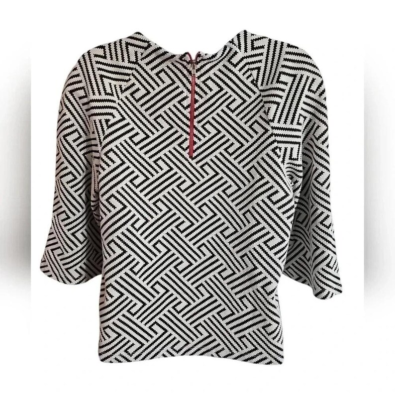 May include: A black and white geometric patterned top with three-quarter sleeves. The top has a high neckline and a red zipper detail. The pattern is a repeating design of black lines on a white background.