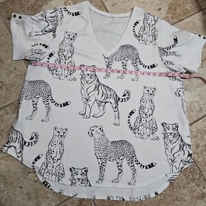 May include: White V-neck shirt with a black and white animal print featuring tigers and cheetahs. The shirt has short sleeves and a curved hem. The shirt measures approximately 24 inches across.