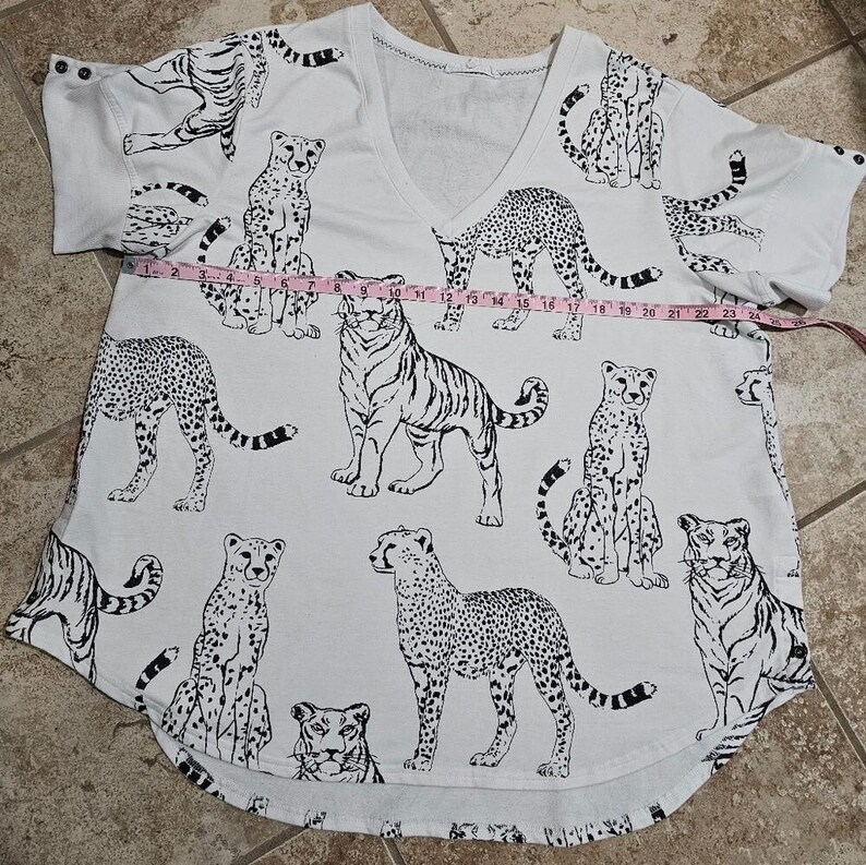 May include: White V-neck shirt with a black and white animal print featuring tigers and cheetahs. The shirt has short sleeves and a curved hem. The shirt measures approximately 24 inches across.