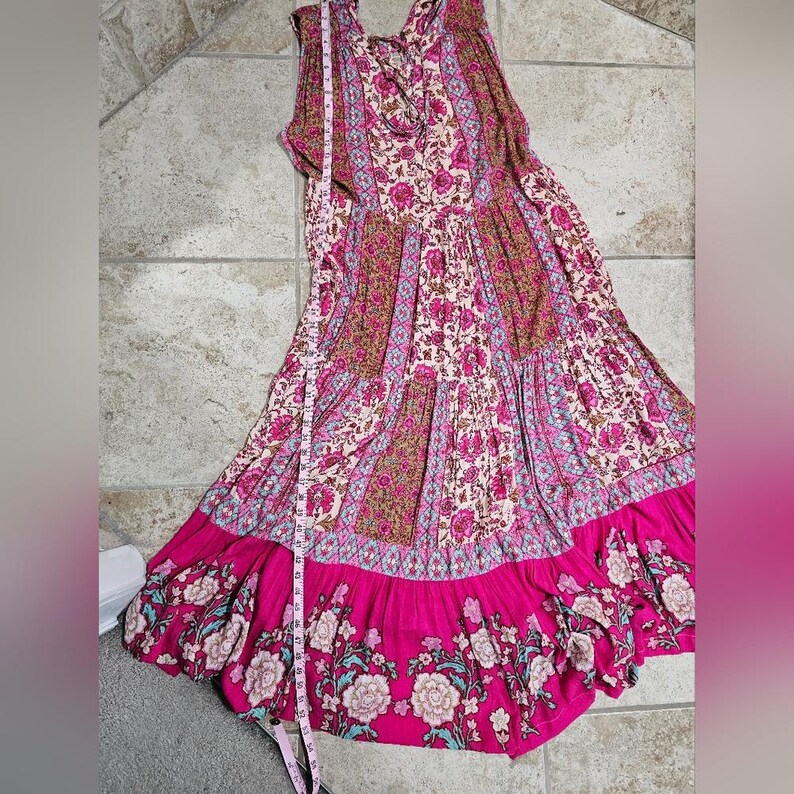 May include: A sleeveless, floor-length dress with a floral print in shades of pink, brown, and cream. The dress features a tiered design with a bright pink hemline and floral border. A measuring tape is next to the dress.