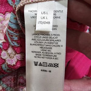 May include: A close-up of a clothing tag with washing instructions in French. The tag includes size information: US L and UK L. The tag also has care symbols for washing, drying, and ironing.