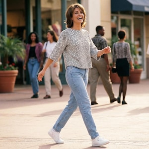 May include: A woman wearing a black and white geometric patterned top, light blue jeans, and white sneakers walks on a paved walkway. Other people are in the background, some wearing jeans and skirts.