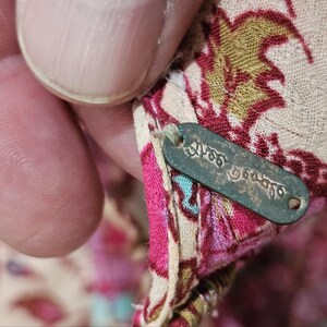 May include: Close-up of a fabric tag with the text "Yves Delorme" in a script font. The tag is attached to a floral patterned fabric with pink, green, and beige hues. The fabric is being held by a person.