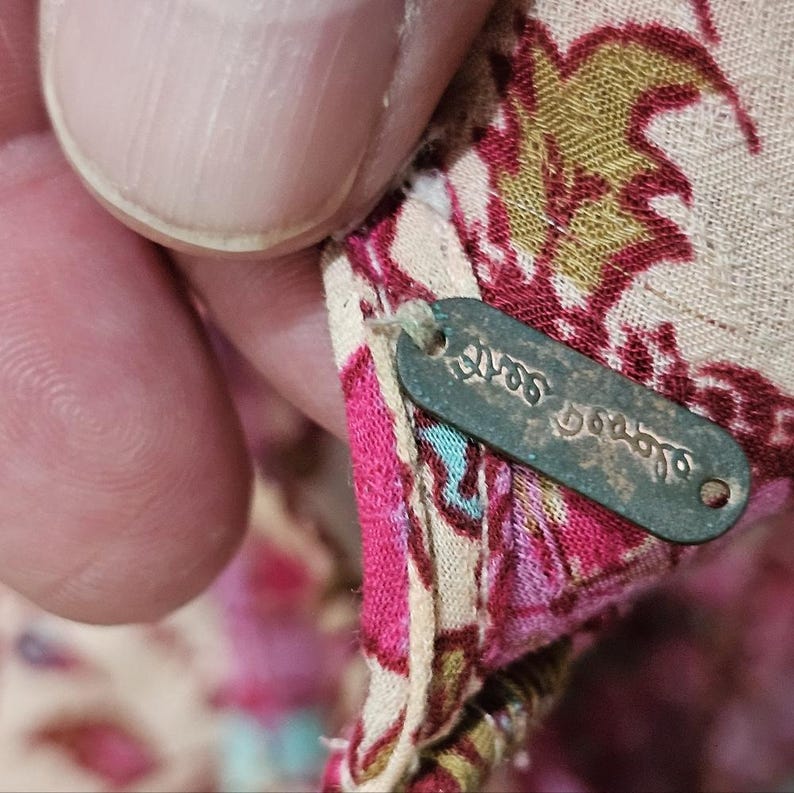 May include: Close-up of a fabric tag with the text "Yves Delorme" in a script font. The tag is attached to a floral patterned fabric with pink, green, and beige hues. The fabric is being held by a person.