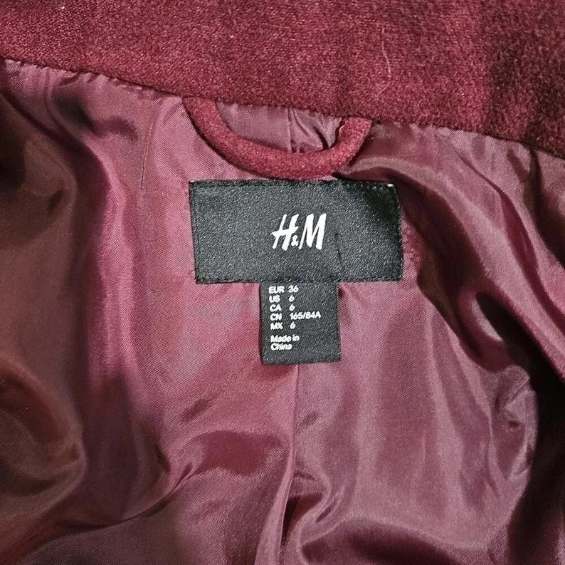 Puede incluir: Primer plano del interior de un abrigo burdeos. El forro es de un tono m&aacute;s claro de burdeos. Una etiqueta negra con el logo de H&M y la informaci&oacute;n de la talla es visible.