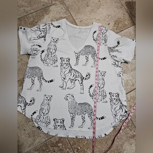 May include: White V-neck t-shirt with a black and white animal print. The print features tigers, cheetahs, and other big cats. The shirt is approximately 29 inches long.