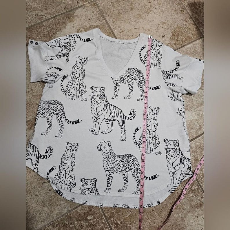 May include: White V-neck t-shirt with a black and white animal print. The print features tigers, cheetahs, and other big cats. The shirt is approximately 29 inches long.