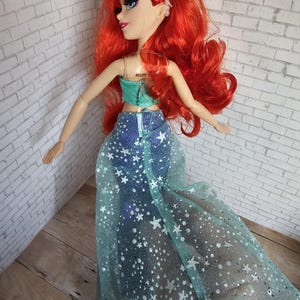 May include: A doll with bright red hair, wearing a teal top and a denim skirt with a sheer, star-patterned overlay. The doll has blue eyes and a purple headband. The doll is posed against a white brick wall.